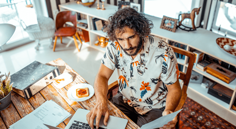 "A man with curly hair and a floral shirt reviewing mortgage rate documents at home, using a laptop with a piece of toast beside him."