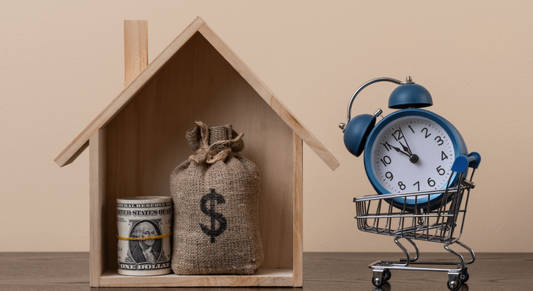 Wooden house structure holding a money bag and rolled cash next to a small shopping cart with a blue alarm clock, symbolizing home value and timing in real estate.